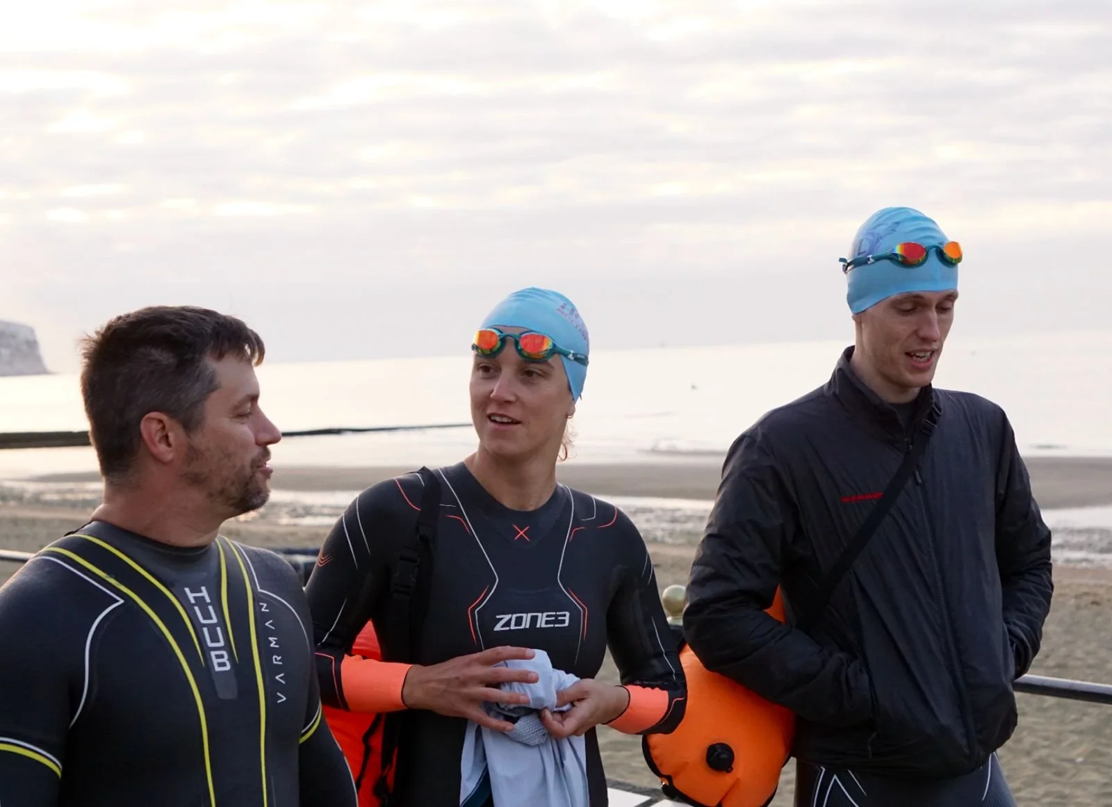 Three swimmers in wetsuits and swim caps with goggles, standing on a beach near the ocean, engaging in conversation during daytime.
