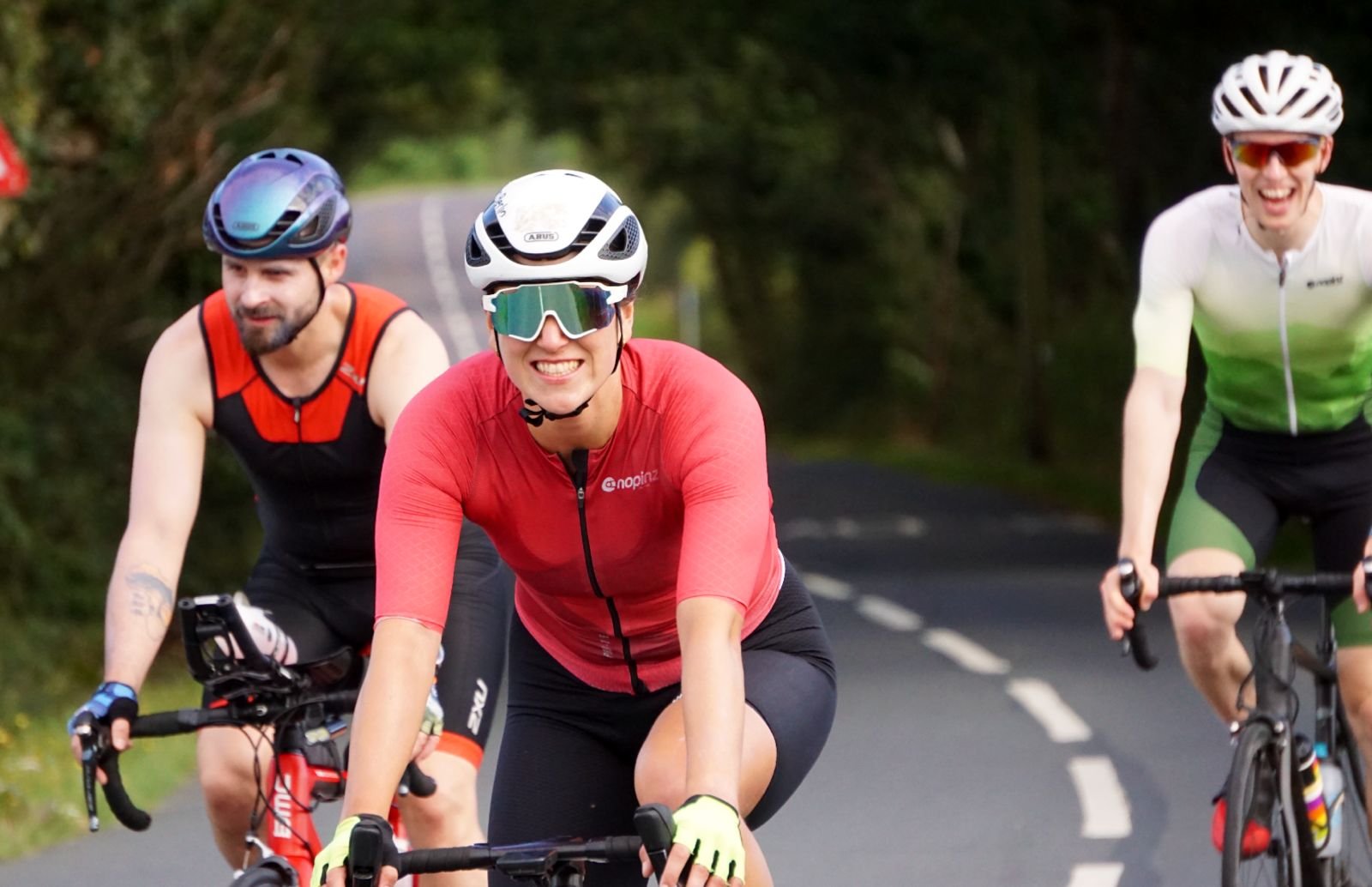 Three cyclists riding on a forested road, smiling and wearing helmets and sportswear.