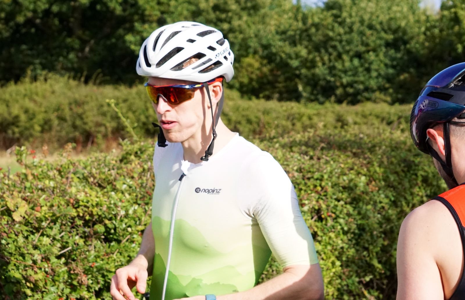 Two men wearing helmets and athletic gear stand outdoors in a green, leafy environment.