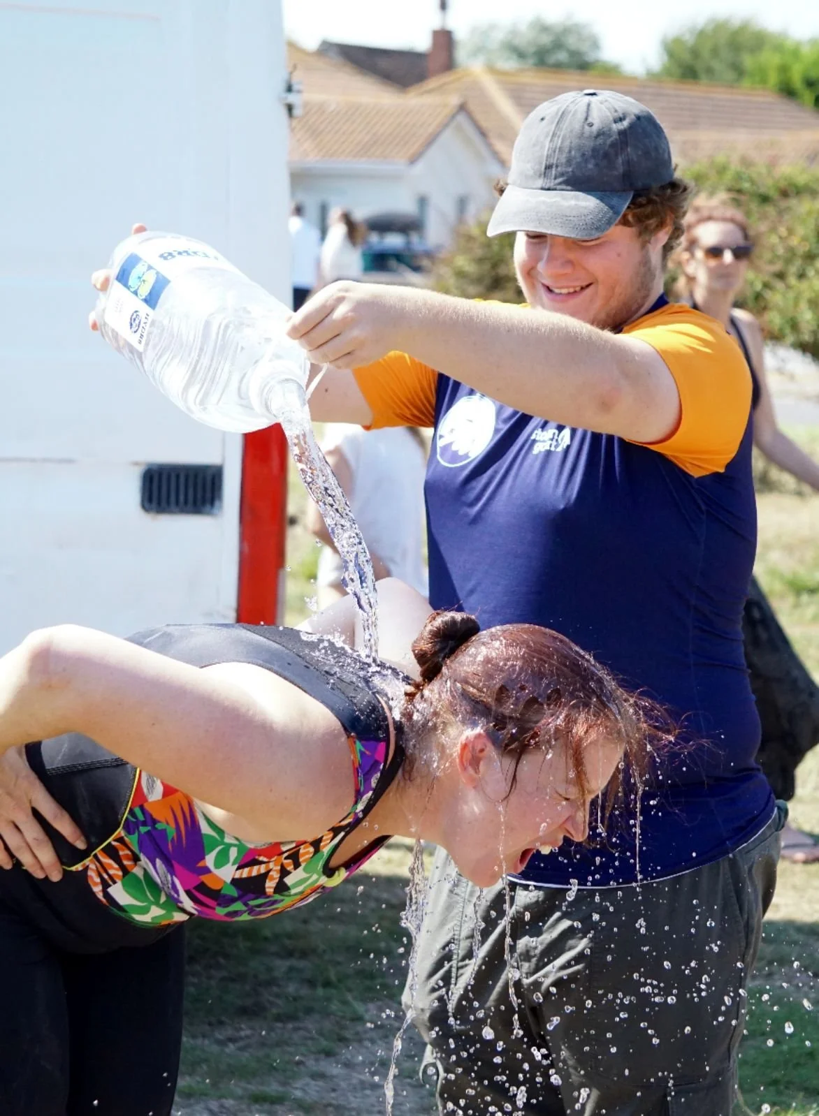 A man pouring water on a woman leaning forward, with water splashing as she closes her eyes to cool off outdoors.