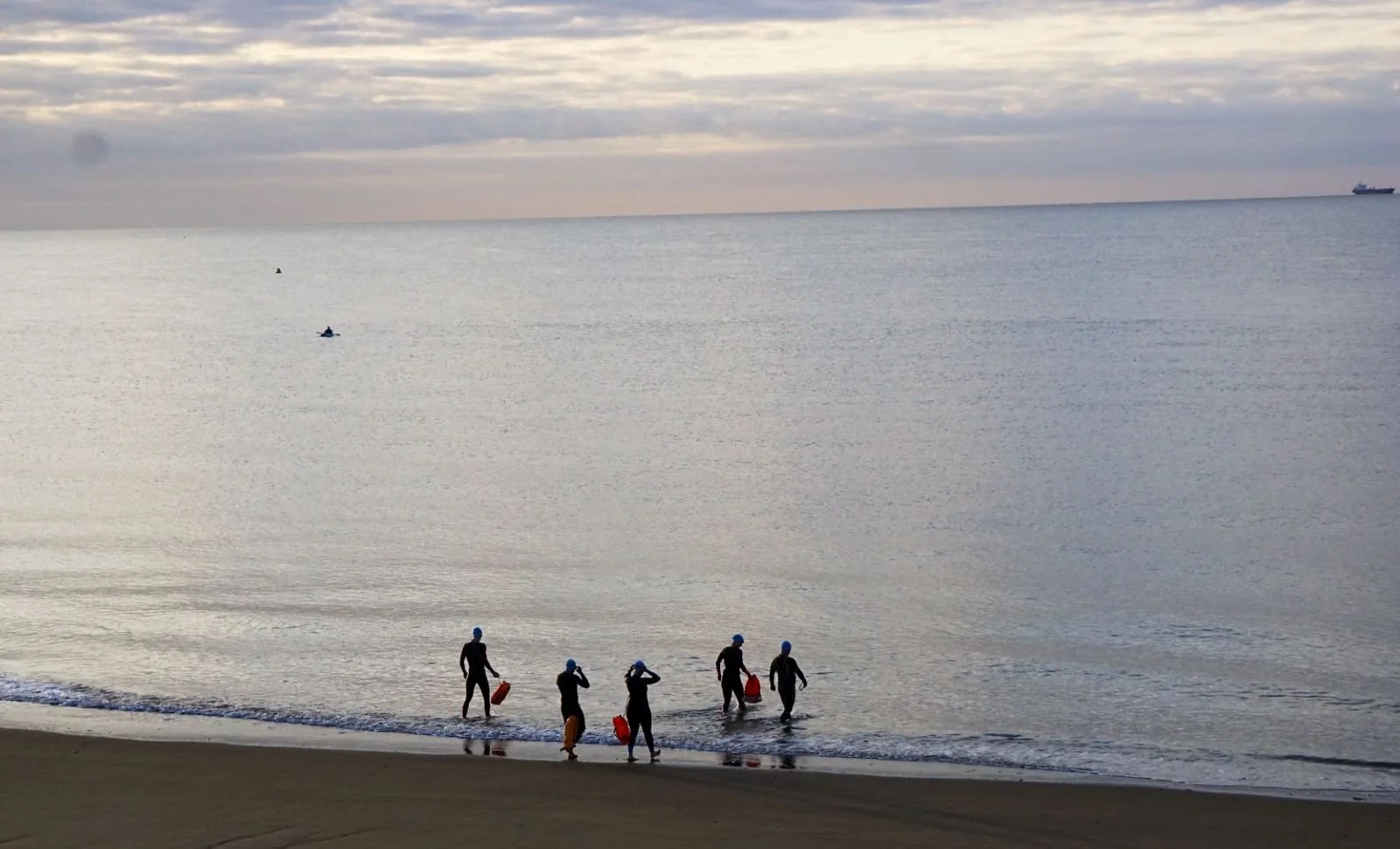 Five people wearing blue swim hats and black wetsuits walking into the ocean carrying orange safety buoys at a beach during sunrise.