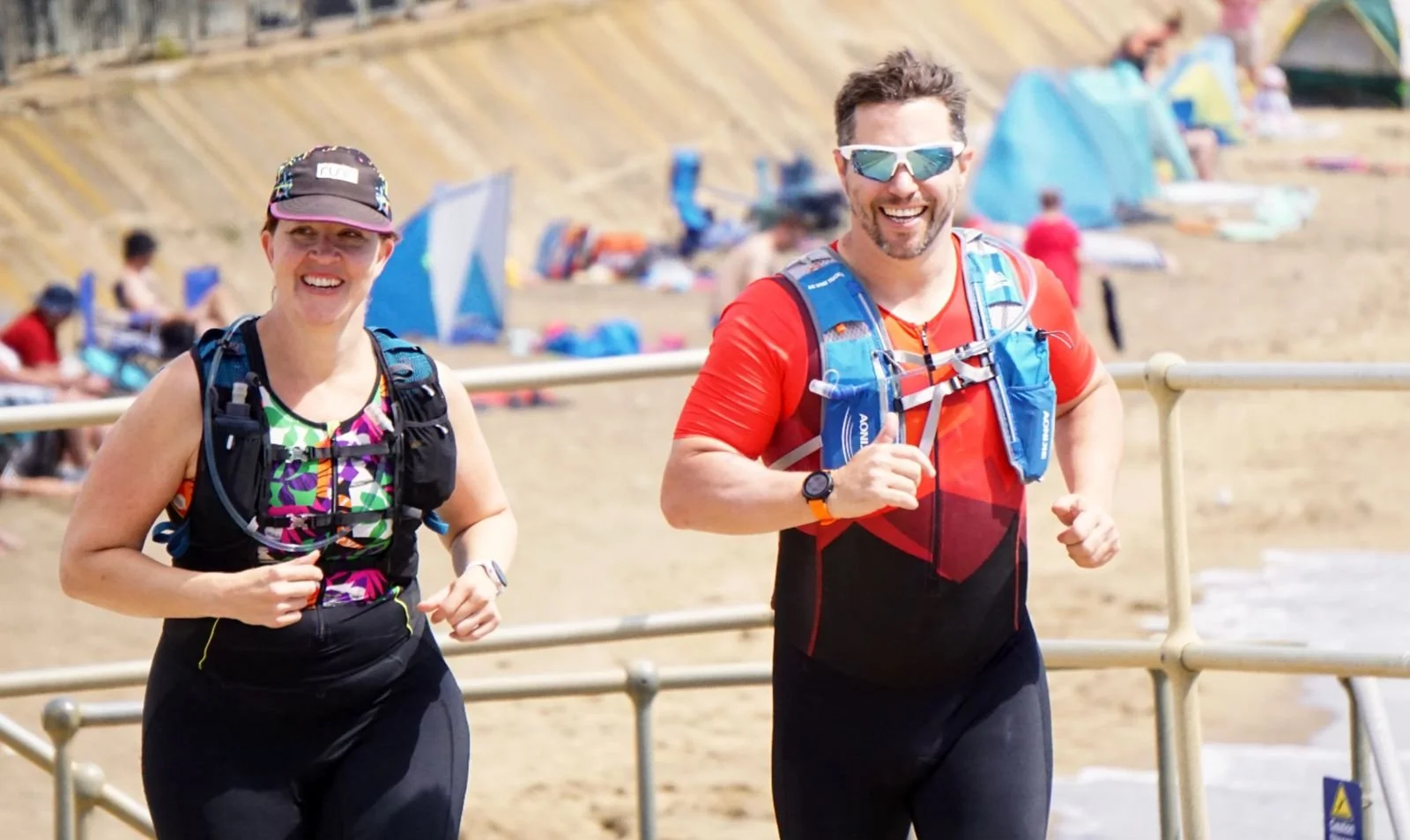 Two people jogging along a boardwalk on a beach, smiling, wearing athletic gear and sunglasses, with tents and beachgoers in the background.