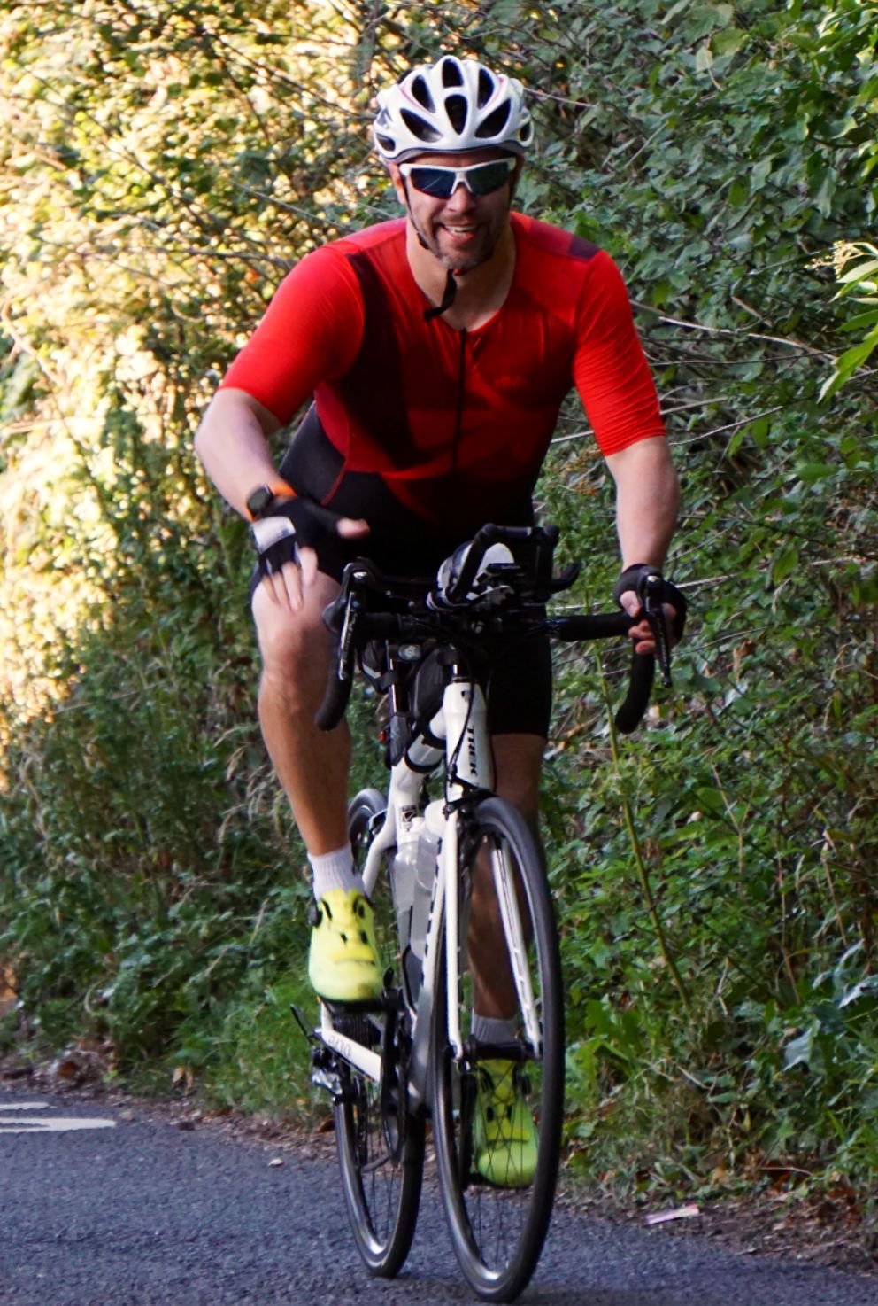 A man riding a bike on a trail through a wooded area, wearing a red and black shirt, yellow shoes, a white helmet, and sunglasses.