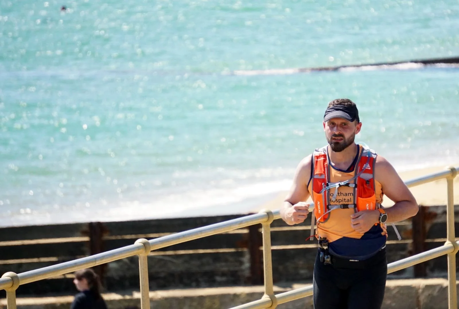 A man running along a waterfront pathway, wearing an orange and gray running vest, black shorts, a black visor, and a watch, with the ocean and a railing behind him.