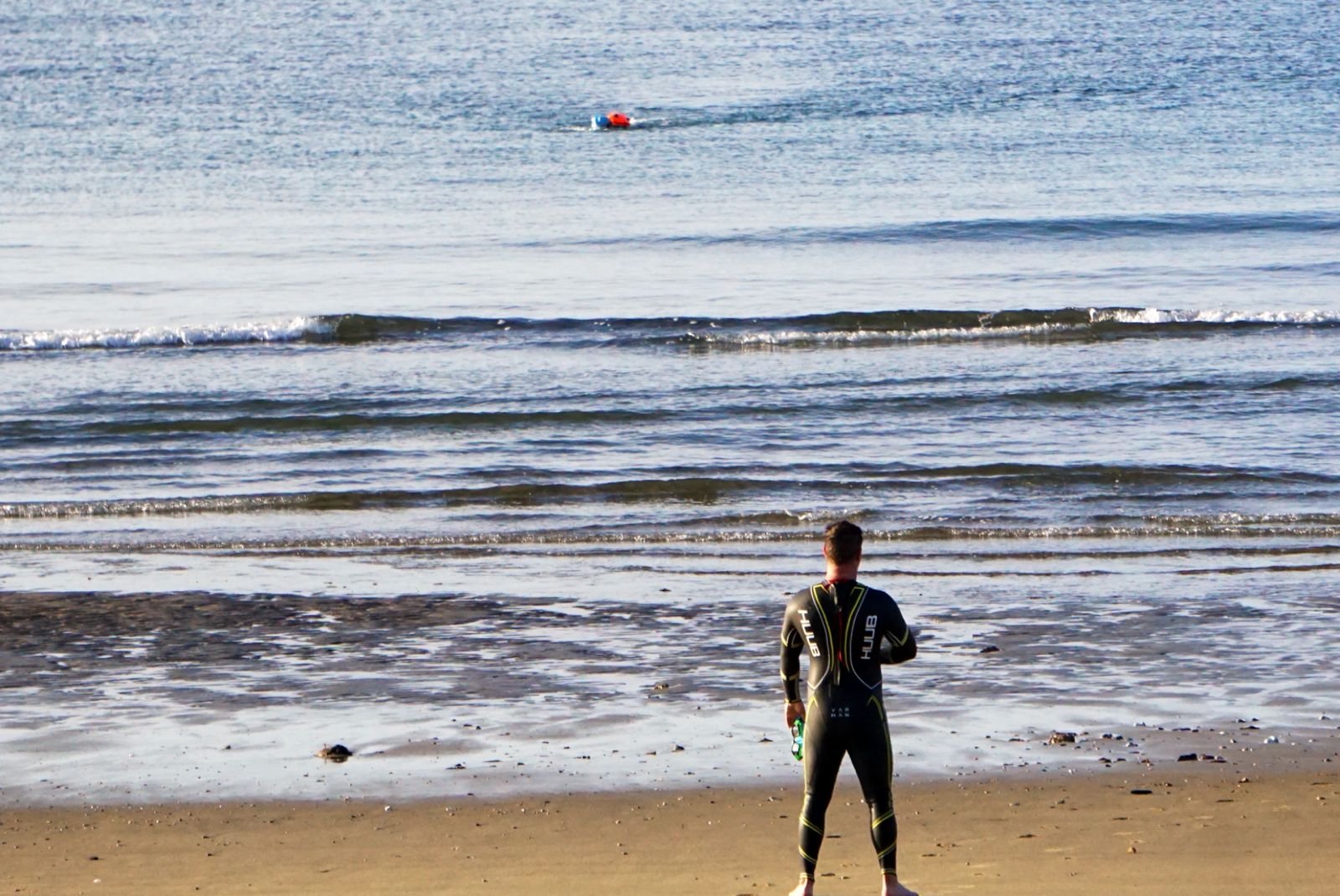 A person in a wetsuit standing on the beach facing the ocean, watching a fellow athlete in the water in the distance.