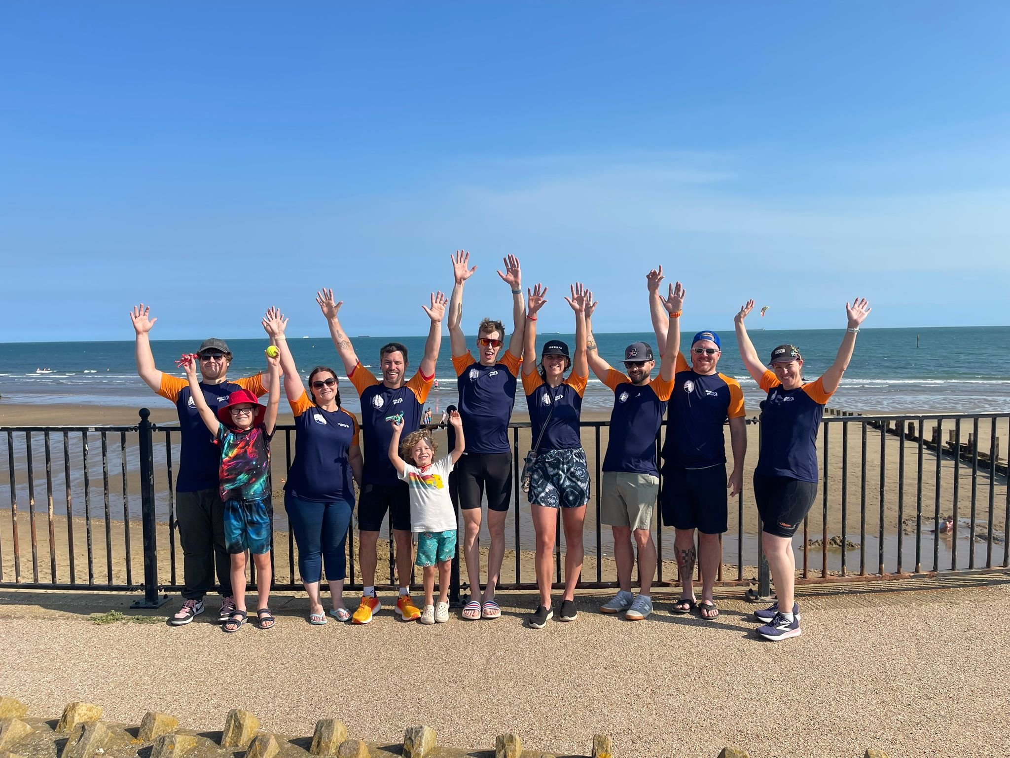 The 2025 RTI team at the beach posing with arms raised, standing near a black fence with the ocean and blue sky in the background.