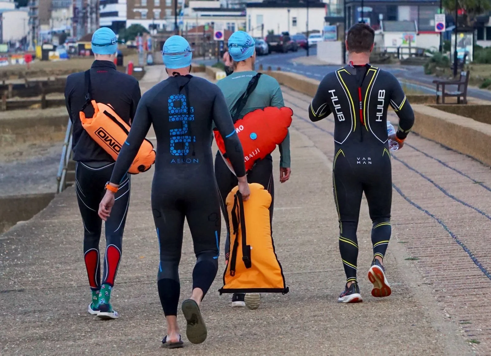 Four athletes walking on a sidewalk, wearing athletic gear and carrying bags and equipment, with a street and buildings in the background.