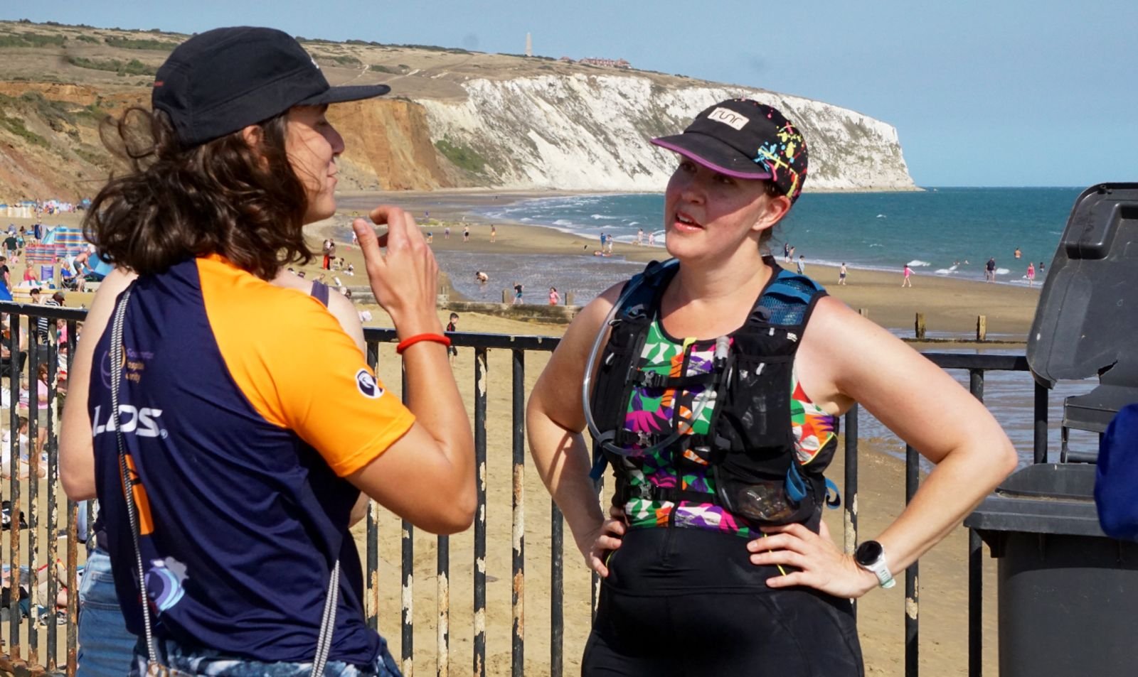 Two women having a conversation at the beach with cliffs and people in the background.