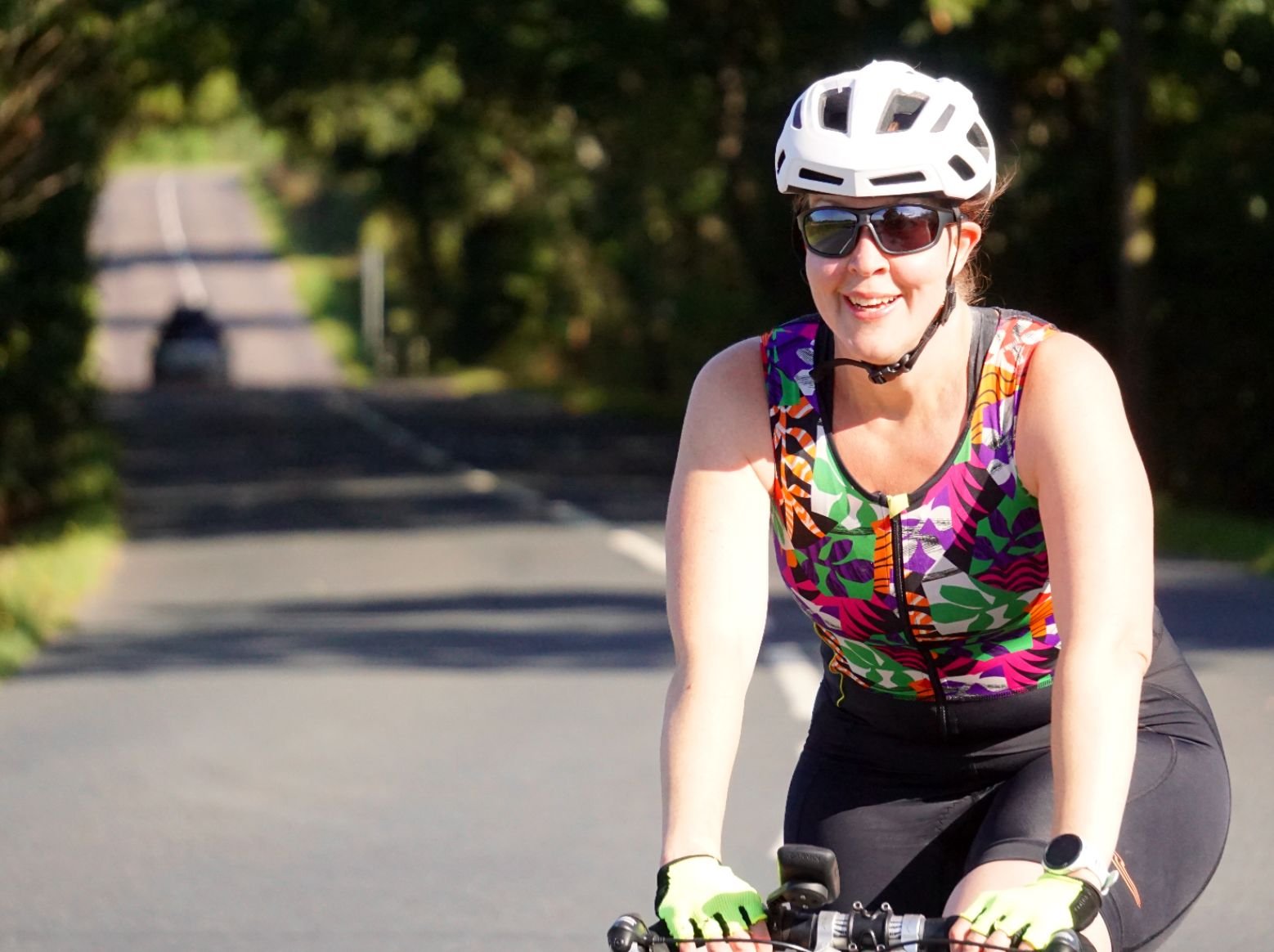 A woman wearing a colorful sleeveless top, sunglasses, helmet, and gloves riding a bicycle on a scenic road surrounded by greenery.