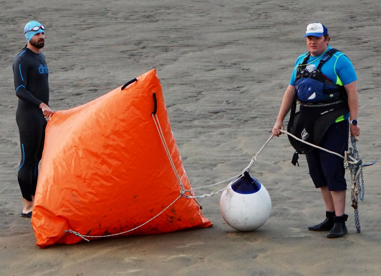 Two men on a sandy beach, one holding an orange flotation device and the other holding a buoy, both dressed in wetsuits and gear.