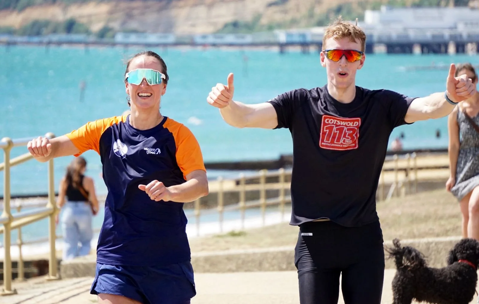 Two runners, a woman and a man, smiling and giving thumbs up while running along a seaside path during a race, with water and a bridge in the background.
