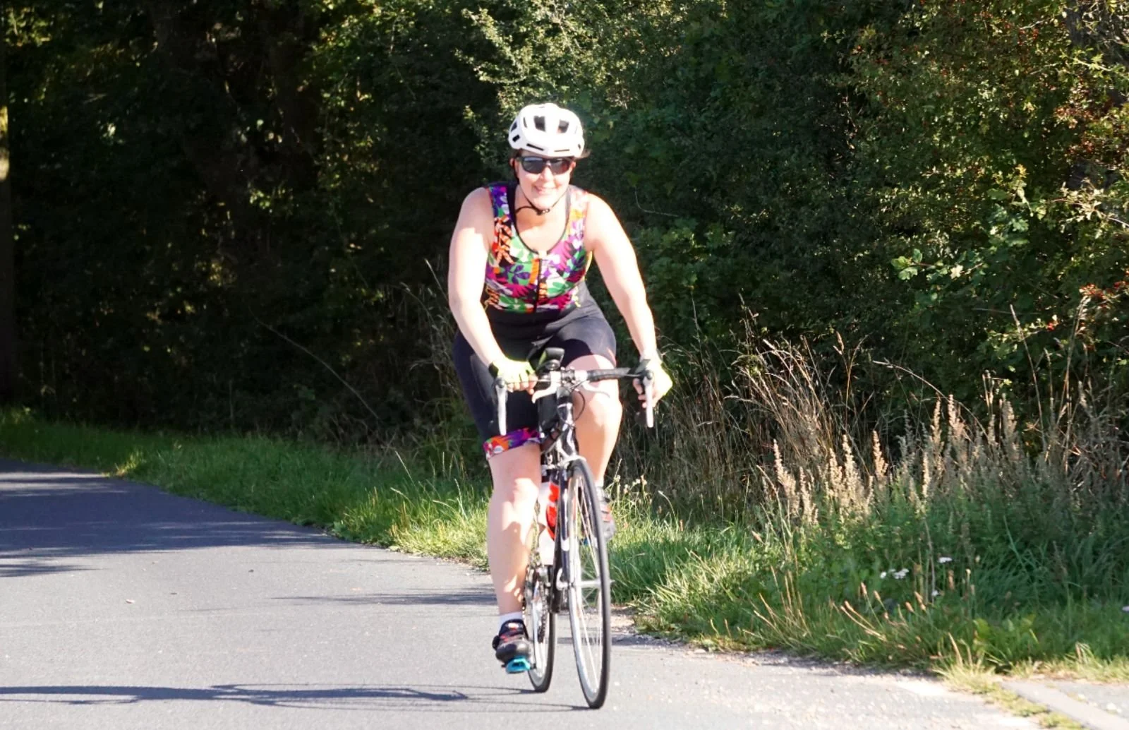 Woman riding a bicycle on a paved road surrounded by greenery, wearing a helmet and sunglasses.