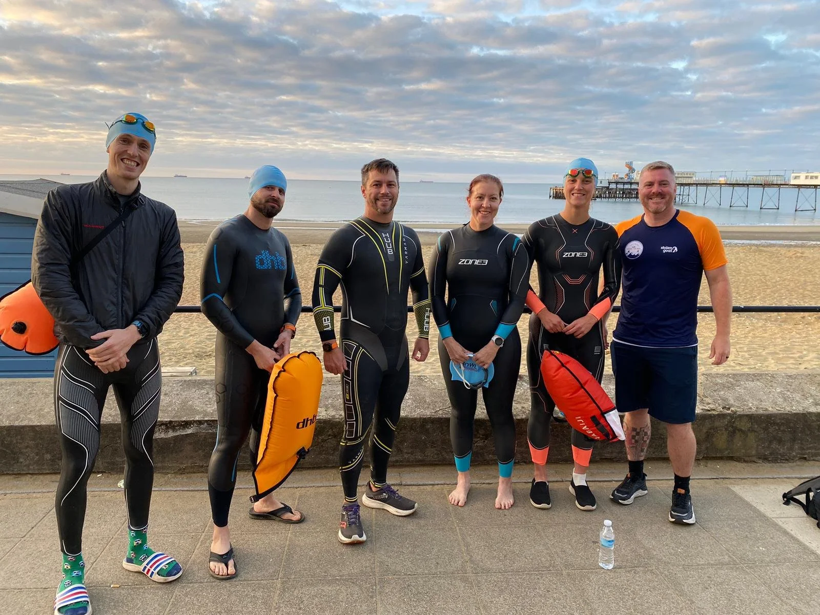 Group of seven people in wetsuits standing on a beach promenade with ocean and cloudy sky in the background.