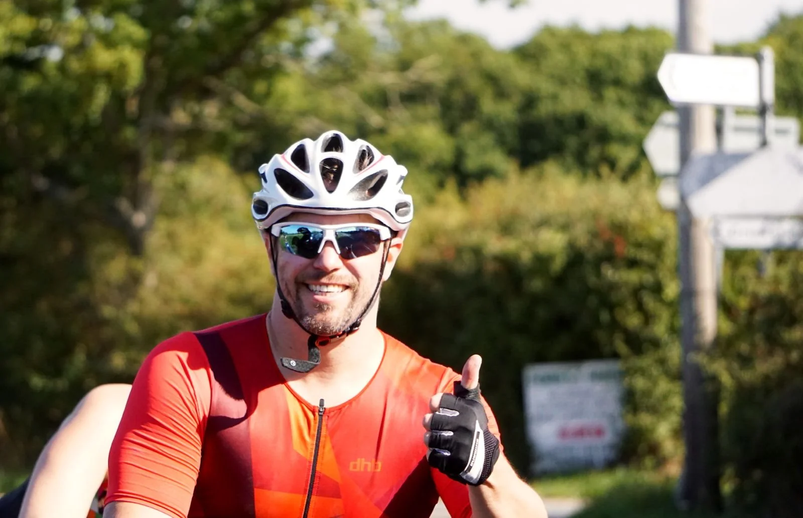 A smiling man wearing a white bike helmet, sunglasses, and an orange and black cycling jersey, giving a thumbs-up gesture during a cycling activity outdoors.