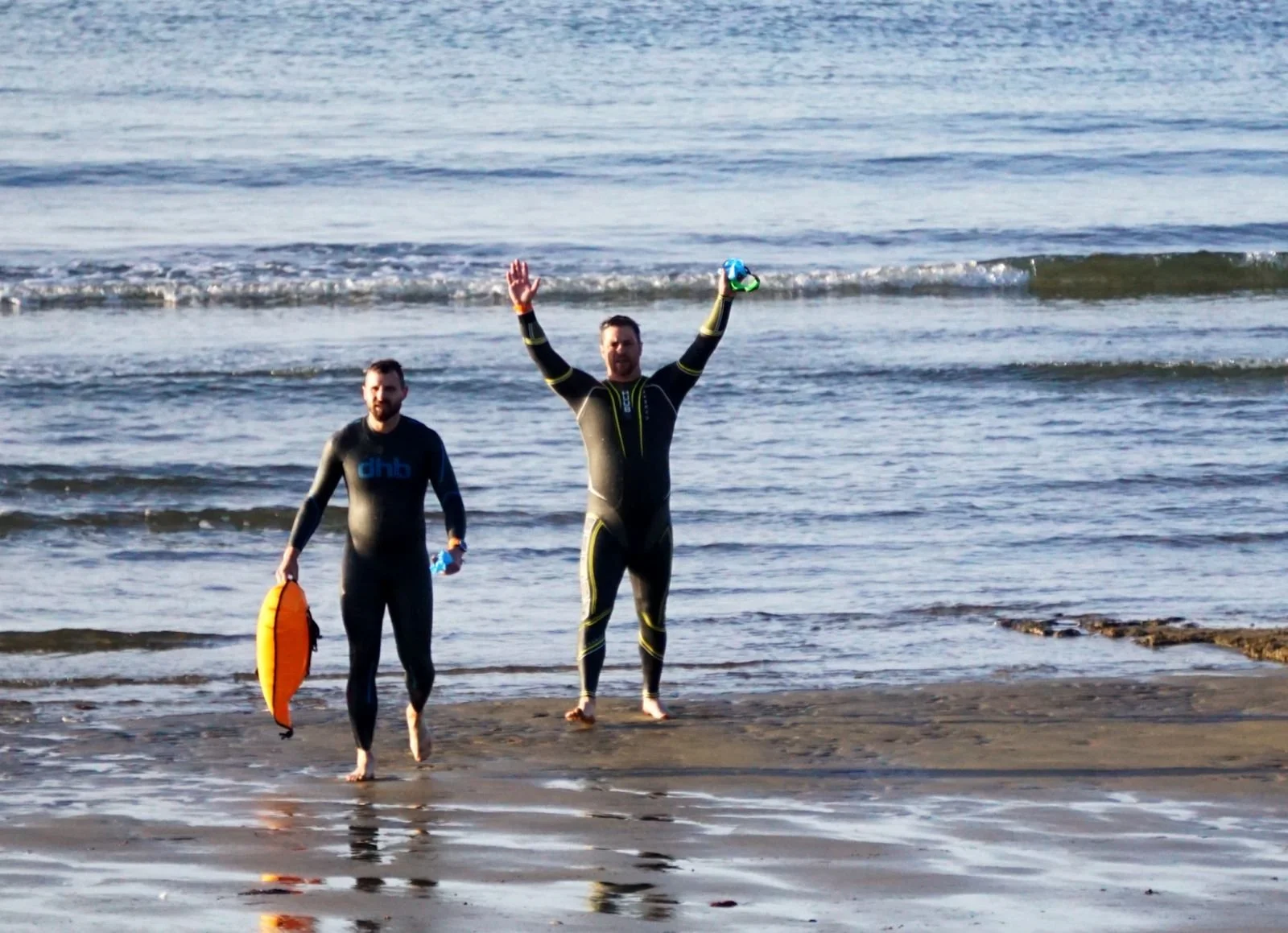 Two people in wetsuits walking out of the ocean, one raising arms in celebration, the other holding a flotation device and a towel, waves in the background.