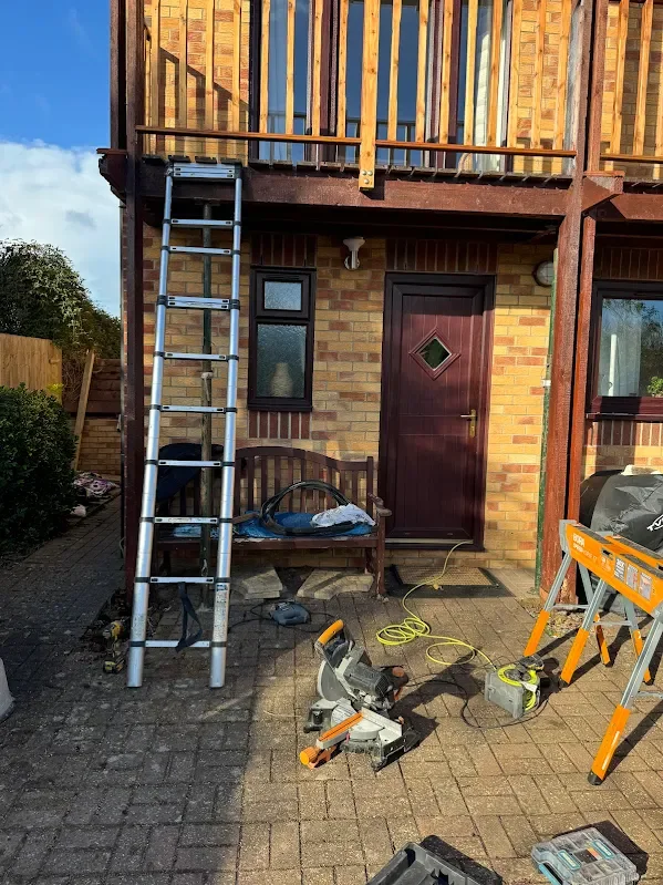 Exterior brick house with door, two small windows, wooden balcony, ladder leaning against the wall, bench with tools, and construction equipment on the ground.