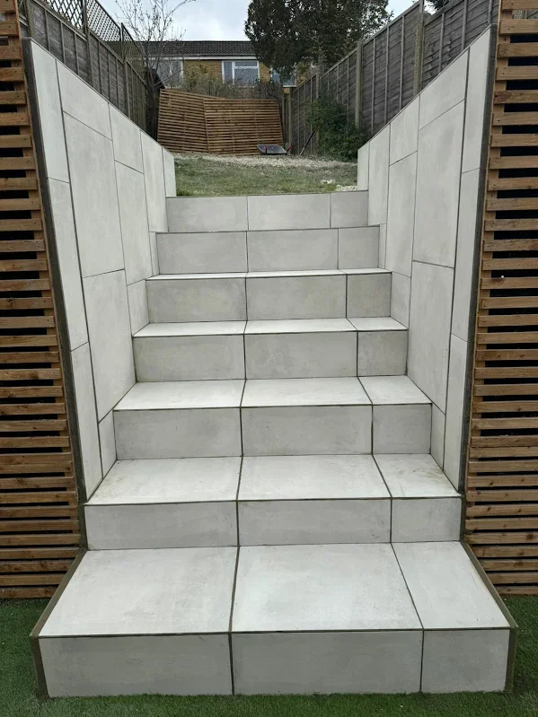 Outdoor staircase with white tile steps and matching tiled walls on each side, leading up to a grassy yard with a wooden fence and garden in the background.