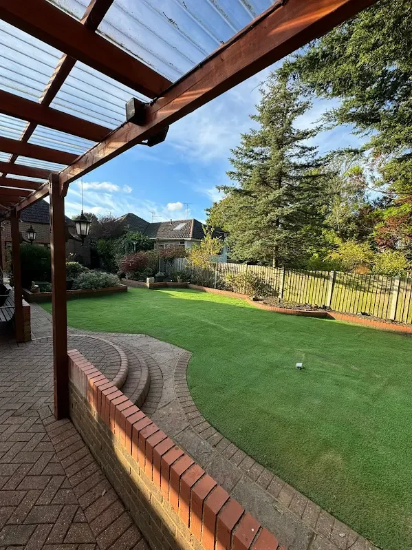 View of a backyard with a patio and a grassy area, viewed from under a partly transparent roof. There are steps leading down to the lawn, a garden bed with plants, and trees along the fence line.