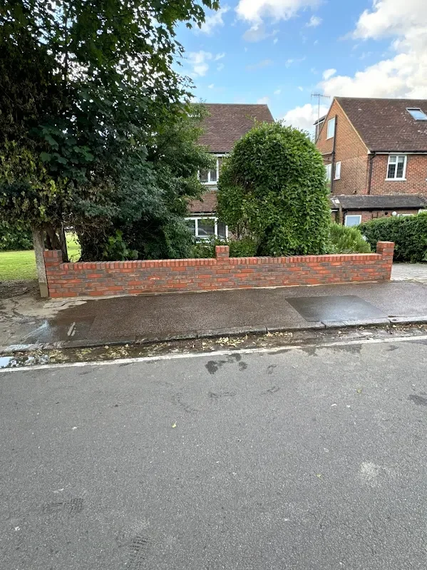 Residential street with a short brick wall, sidewalk, and houses in the background, under a partly cloudy sky.