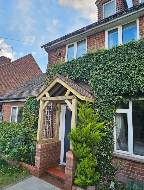 A brick house with green vines covering part of the exterior, a small front porch with a wooden frame and a shingled roof, and a small plant next to the porch. There is a bay window on the right side and other houses nearby.