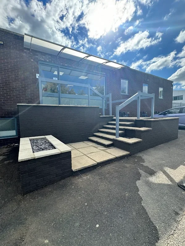 Exterior view of a modern brick building with a raised outdoor patio area, stairs, and glass railings, under partly cloudy skies.