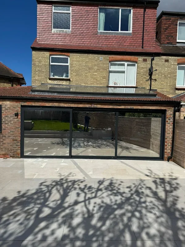 A brick house with large glass sliding doors leading to a patio. The house has multiple windows, some with curtains. There is a potted plant and a person reflected in the glass. Shadows of trees are cast on the tiled patio surface.