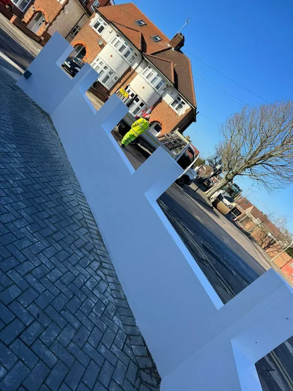 Large white decorative installation with vertical slats on a paved sidewalk, with a worker in a high-visibility jacket working nearby and buildings in the background.