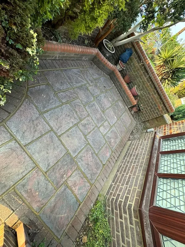 A small backyard patio with stone paving, bordered by brick and plants, with garden tools and potted plants along the wall.