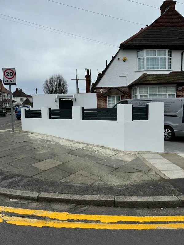 White wall with black horizontal slats and a house behind it, located at the corner of a street with a 20 mph zone sign.