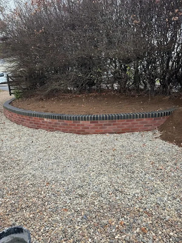 A low curved brick wall with black brick top, separating a dirt area with leafless bushes from a gravel ground.