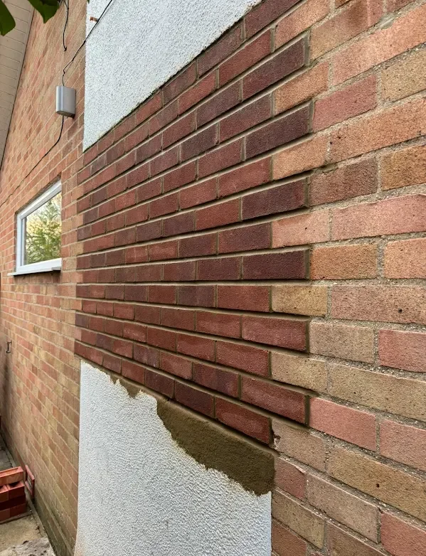 A brick wall being repaired, with new bricks and mortar applied on a section of the wall. There is a window and a small vent on the wall.