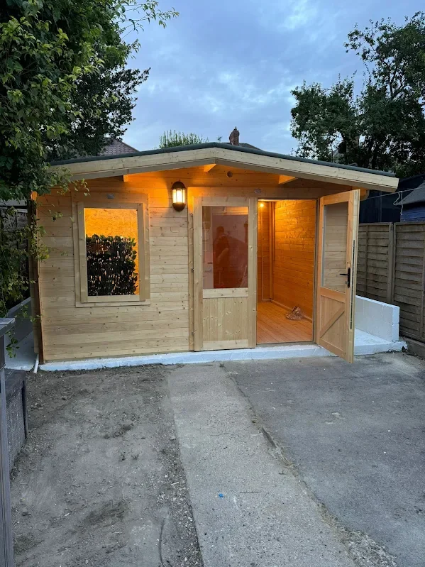 Front view of a small wooden shed with a door and window, illuminated by a porch light, situated on a concrete foundation, with trees and a fence in the background at dusk.