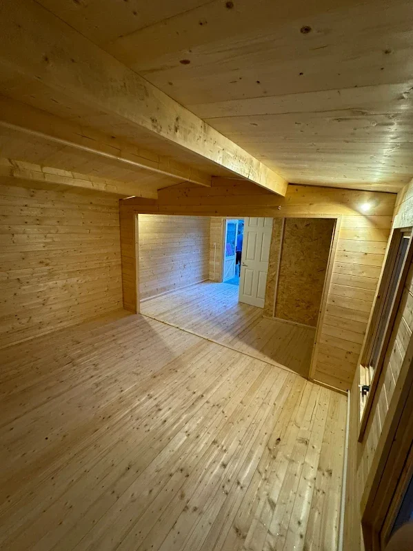 Unfinished interior of a wooden room with a doorway and windows, featuring natural wood paneling on floors, walls, and ceiling.