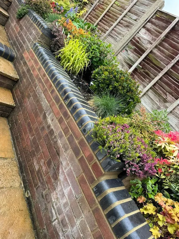 A brick garden bed with colorful flowers and plants, separated from a paved pathway by a curved black and yellow brick border, with a wooden fence in the background.