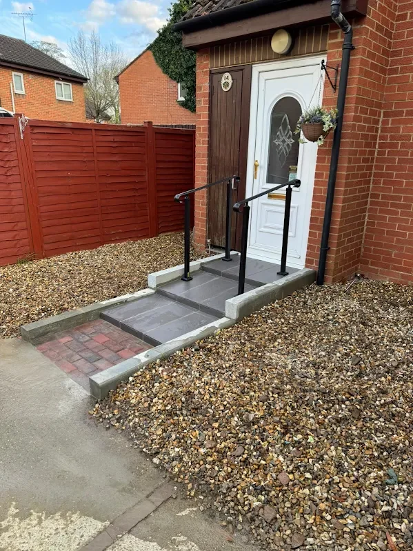 Entrance to a house with a ramp for wheelchair access, made of gray tiles and bordered by concrete, with a white door and black handrails on both sides. Surrounding the entrance is a gravel yard with a red wooden fence.