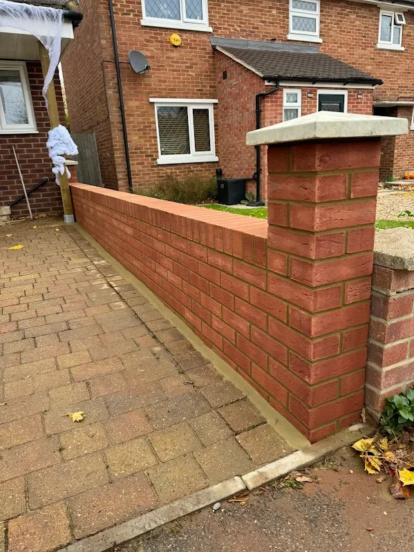 Red brick wall with concrete cap at sidewalk boundary in front of residential brick house with white window frames and satellite dish.
