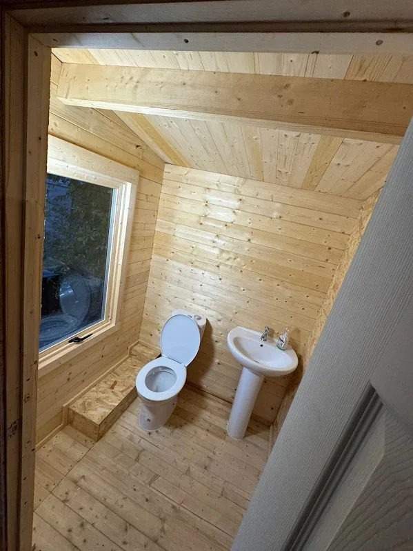 A small bathroom with wood-paneled walls and ceiling, featuring a toilet with the lid open and a pedestal sink. There is a large window on the left side bringing in natural light.