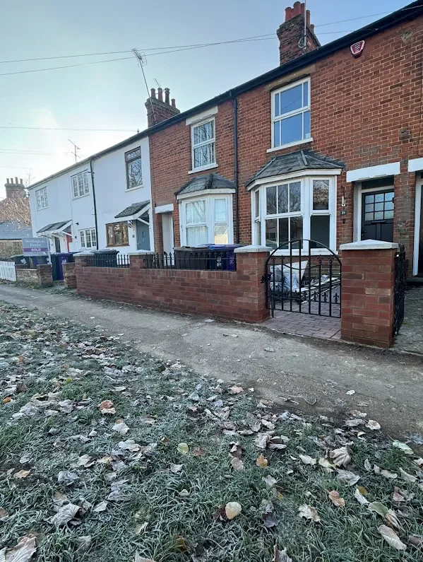 A row of brick houses with small front gardens and metal gates, situated along a narrow dirt sidewalk with fallen leaves.