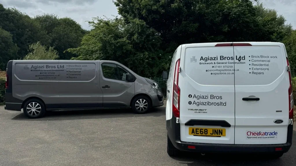 Two vans parked on a paved surface with a background of trees and an overcast sky. The van on the left is gray with signage for Agiazi Bros Ltd, a brickwork and general construction company. The van on the right is white with similar signage, including contact information and services offered, and has a UK license plate reading AE68 JNN.