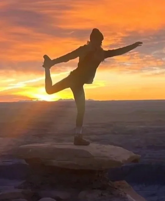 Person performing a yoga pose on a rock at sunset by the water.