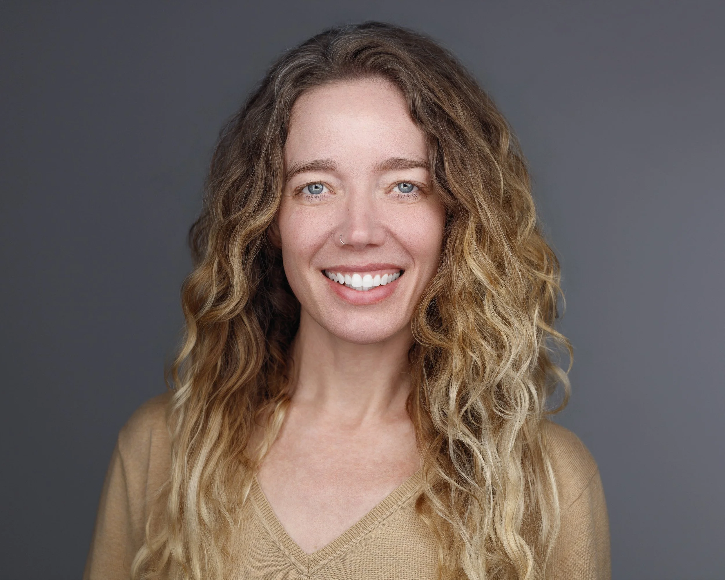 A smiling woman with curly blonde hair, blue eyes, and a nose ring, wearing a beige sweater, against a gray background.