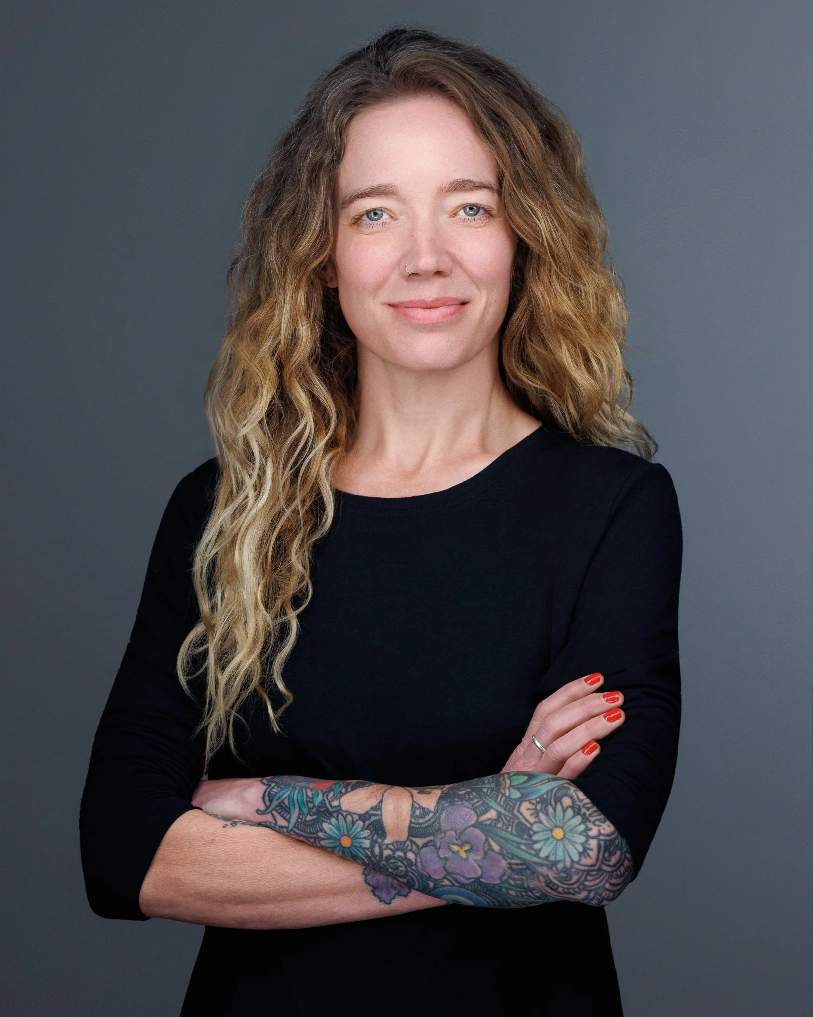 A woman with long curly hair and blue eyes, wearing a black top, with colorful tattoos on her forearm, and red nail polish, standing with arms crossed against a gray background.