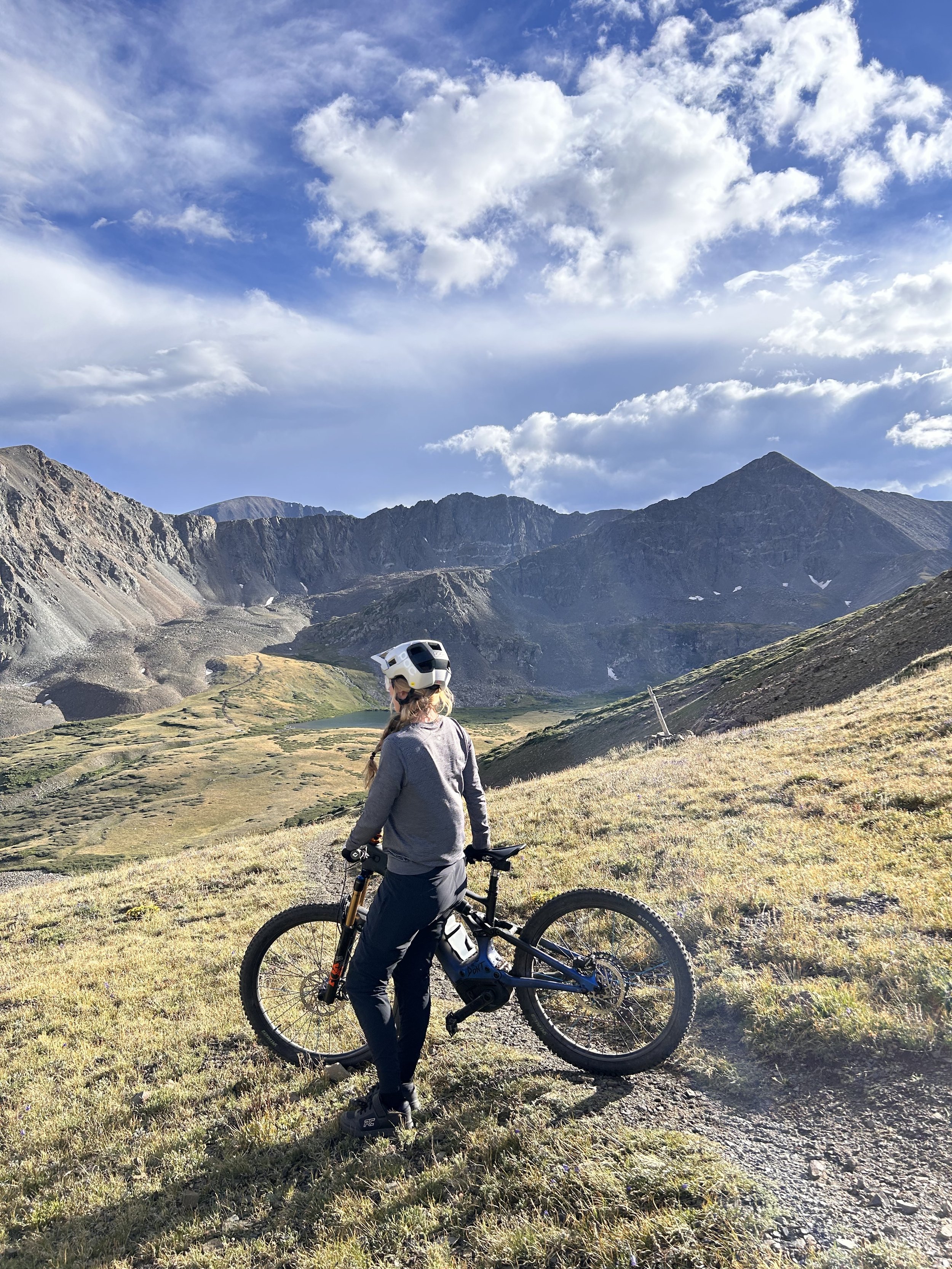 A woman wearing a helmet standing next to a mountain bike on a grassy trail in a mountainous landscape under a partly cloudy sky.