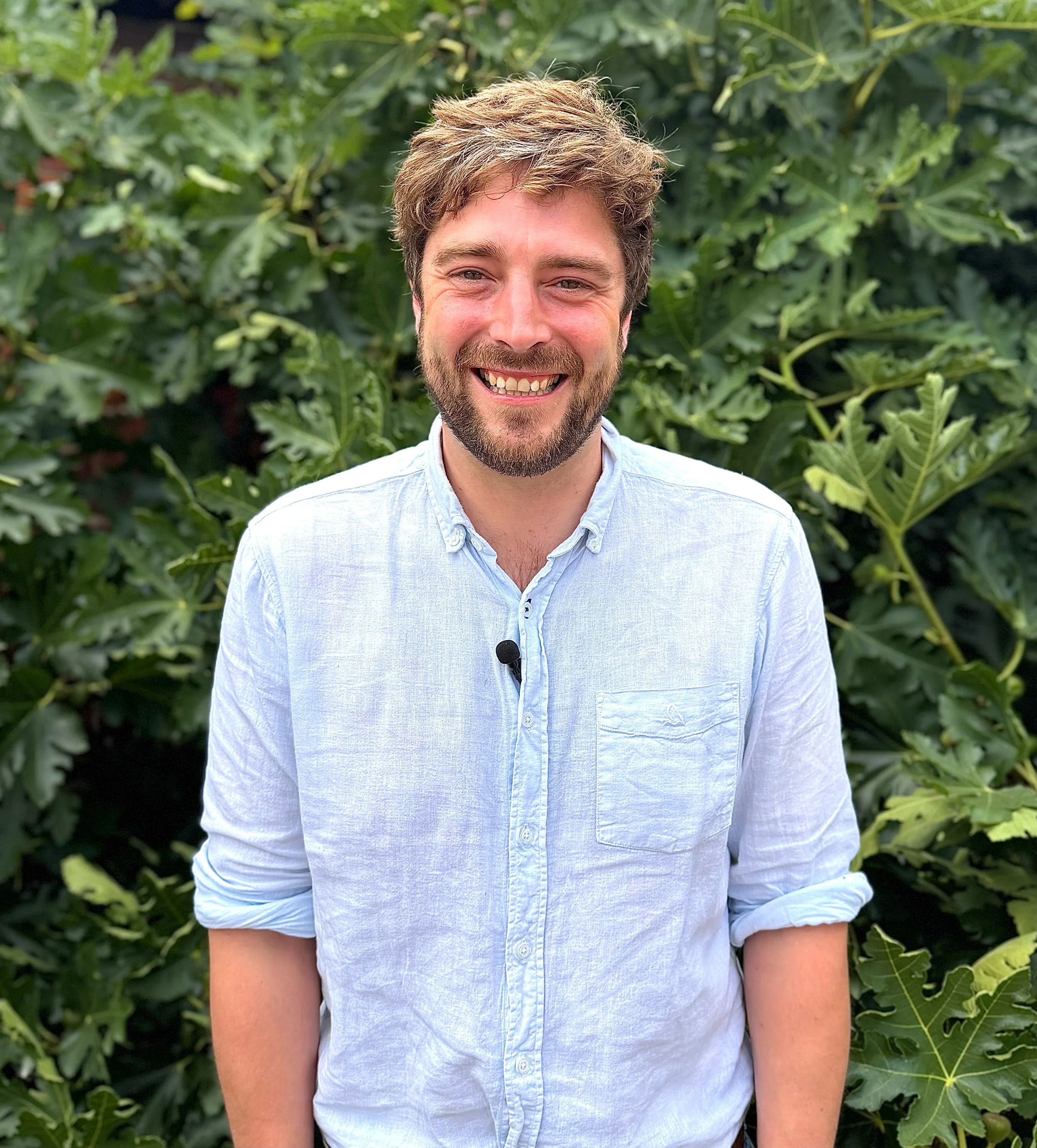 A young male chef wearing a white chef jacket, standing outdoors near a body of water during sunset with a city skyline in the background. He has short brown hair and is crossing his arms, looking confidently at the camera.