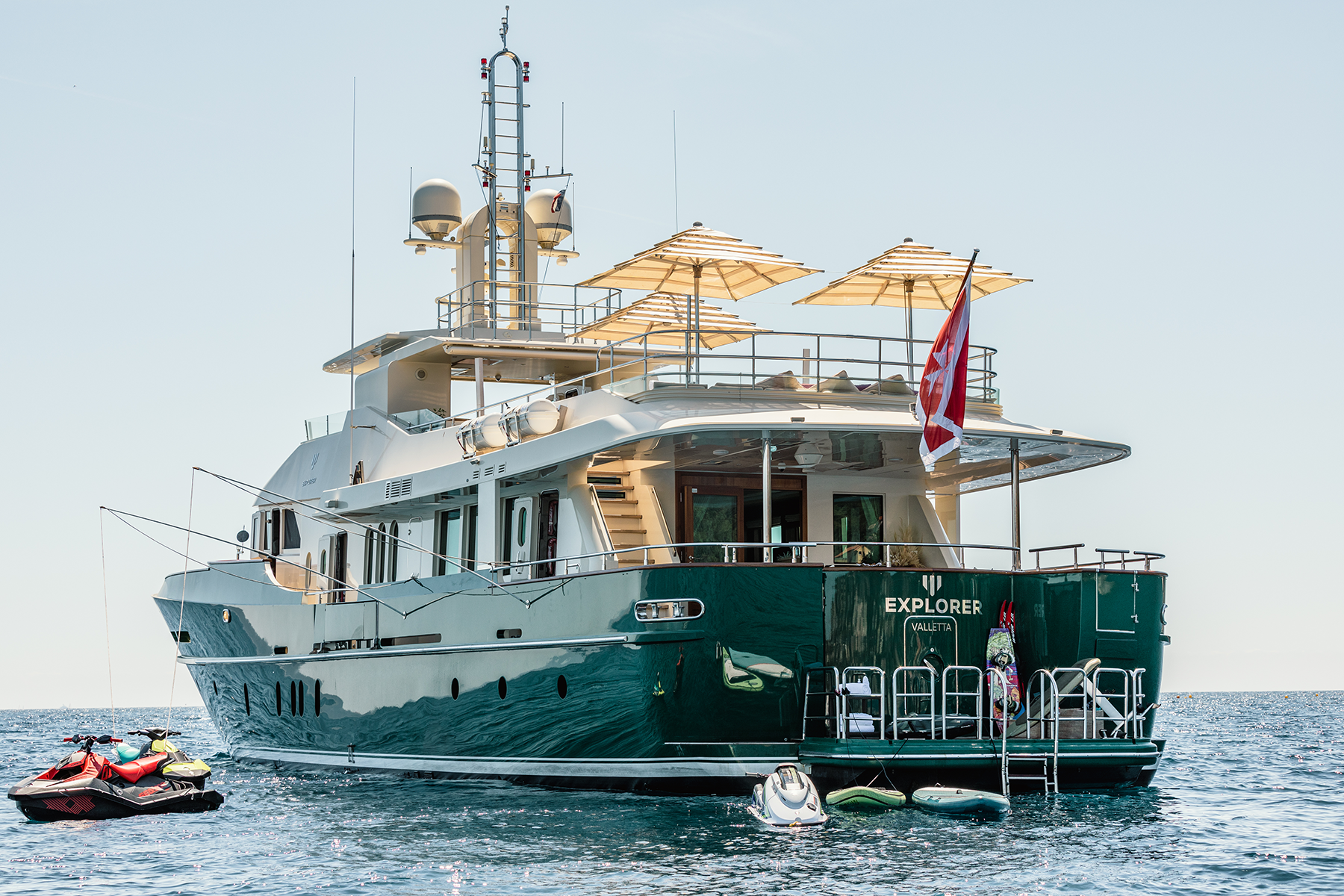 A luxury yacht named 'Explorer Valletta' docked at sea with several jet skis and small boats nearby, light-colored umbrellas on the upper deck, and a flag flying at the stern.