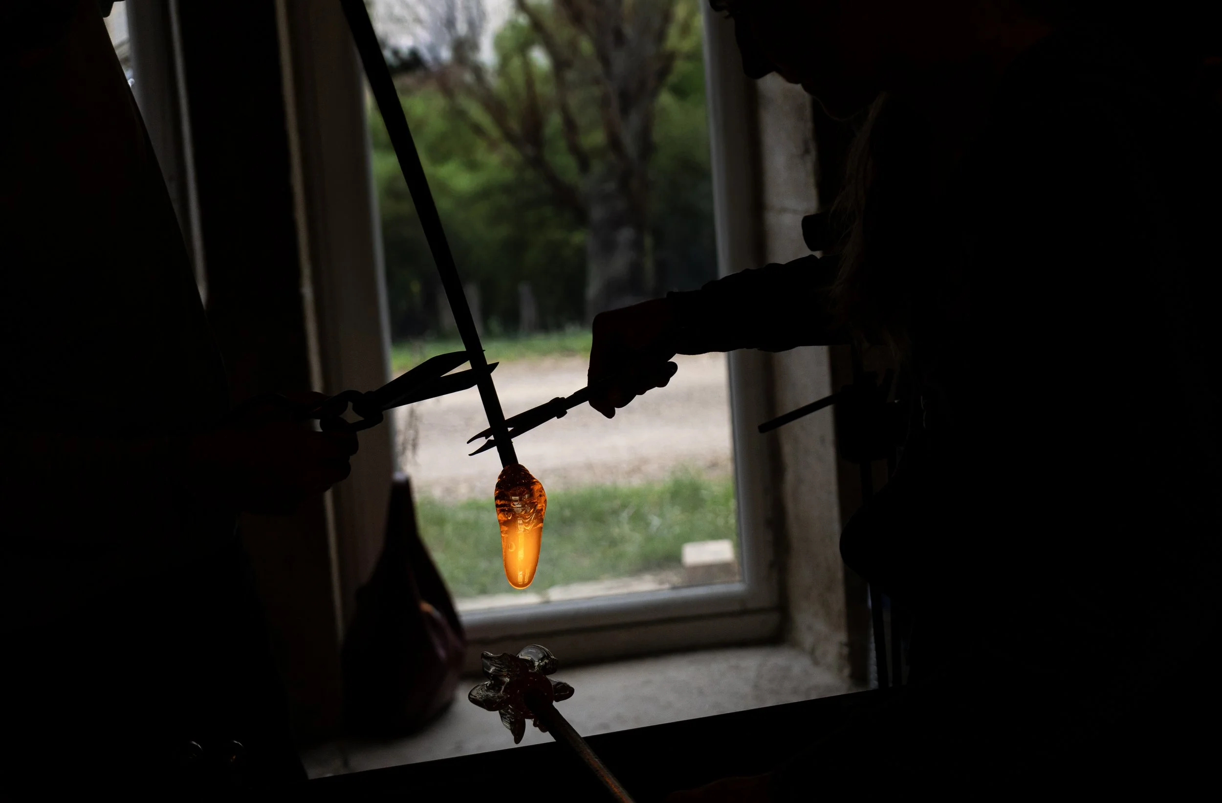 Photographie de deux artistes verriers soufflent du verre chaud dans un atelier, processus visible par la silhouette contre la lumière d'une fenêtre, avec un décor extérieur verdoyant.