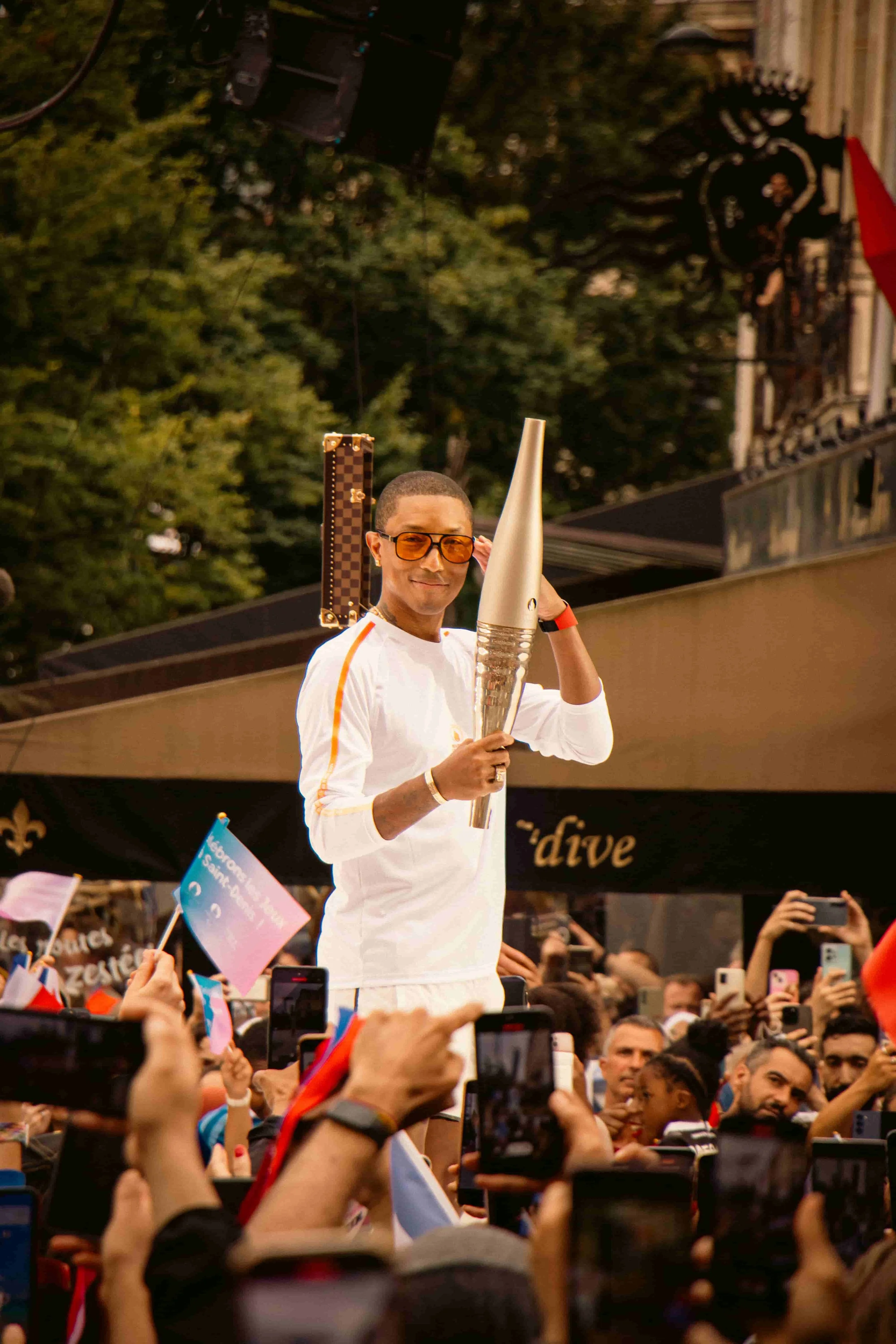 Photographie du chanteur Pharrell Williams à la Basilique Saint-Denis, portant le dernier relai de la flamme olympique en 2024. Il porte des lunettes orange lors d'un événement avec beaucoup de spectateurs prenant des photos. 
