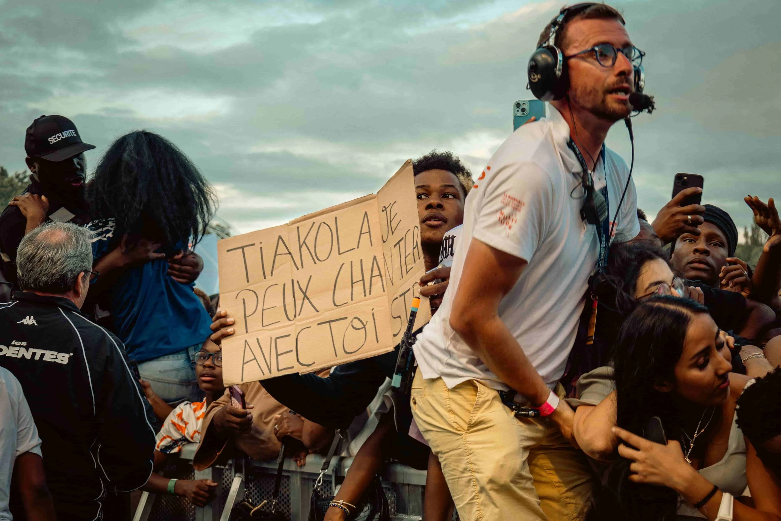 Photographie de la foule lors du concert de Tiakola. Un homme tient une pancarte en français qui dit "Tiakola, je peux chanter avec toi". Plusieurs personnes sont visibles, certaines utilisant un téléphone, et l'ambiance paraît intense et engagée.