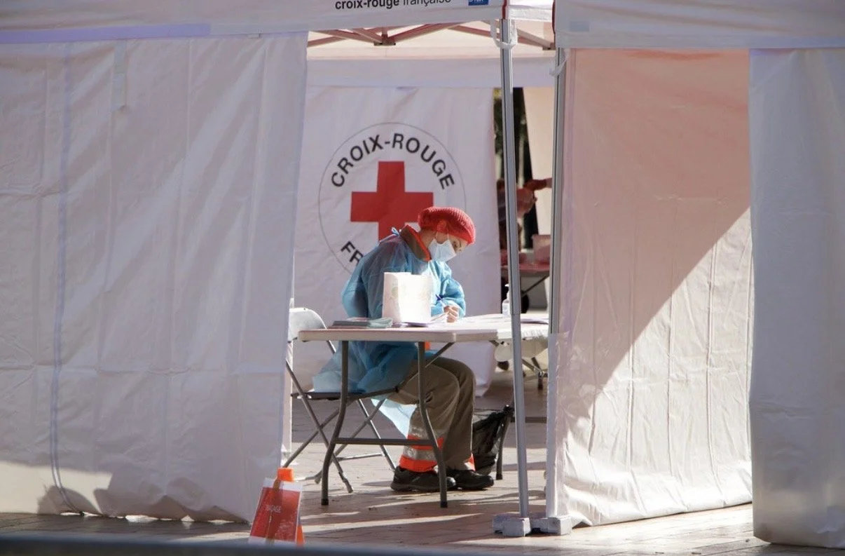 Photographie d'une personne en équipement de protection, assise à une table dans une tente placée à Dijon, à république. Elle travaille pour le centre de dépistage itinérant du COVID-19 de la Croix-Rouge française.