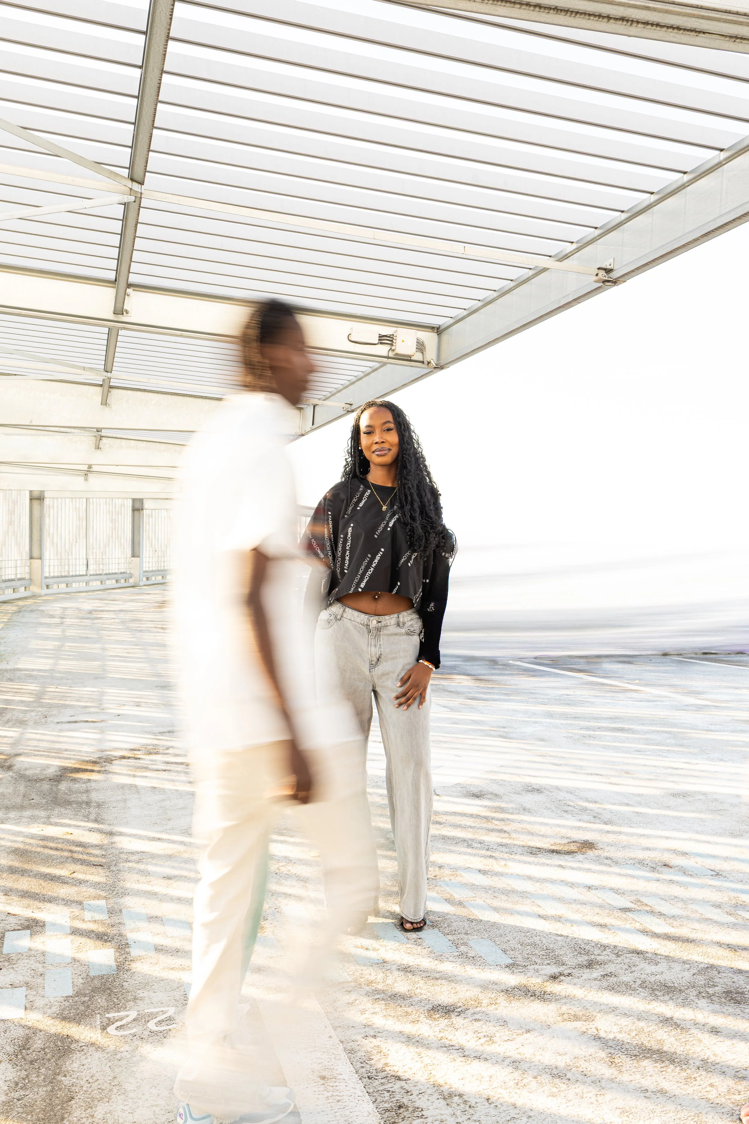  Photographie de mode d'une femme et d'un homme. La femme en arrière-plan porte un crop top noir avec un motif blanc et a de longs cheveux bouclés noirs. L'homme en premier plan est floue, avec des vêtements clairs africains. 